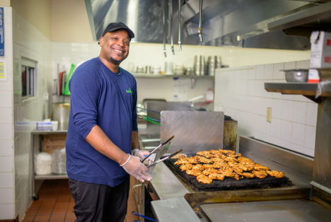 A man with a hat smiles at the camera holding a pair of kitchen thongs. He is standing next to a stove top with food that is cooking on the stove top.