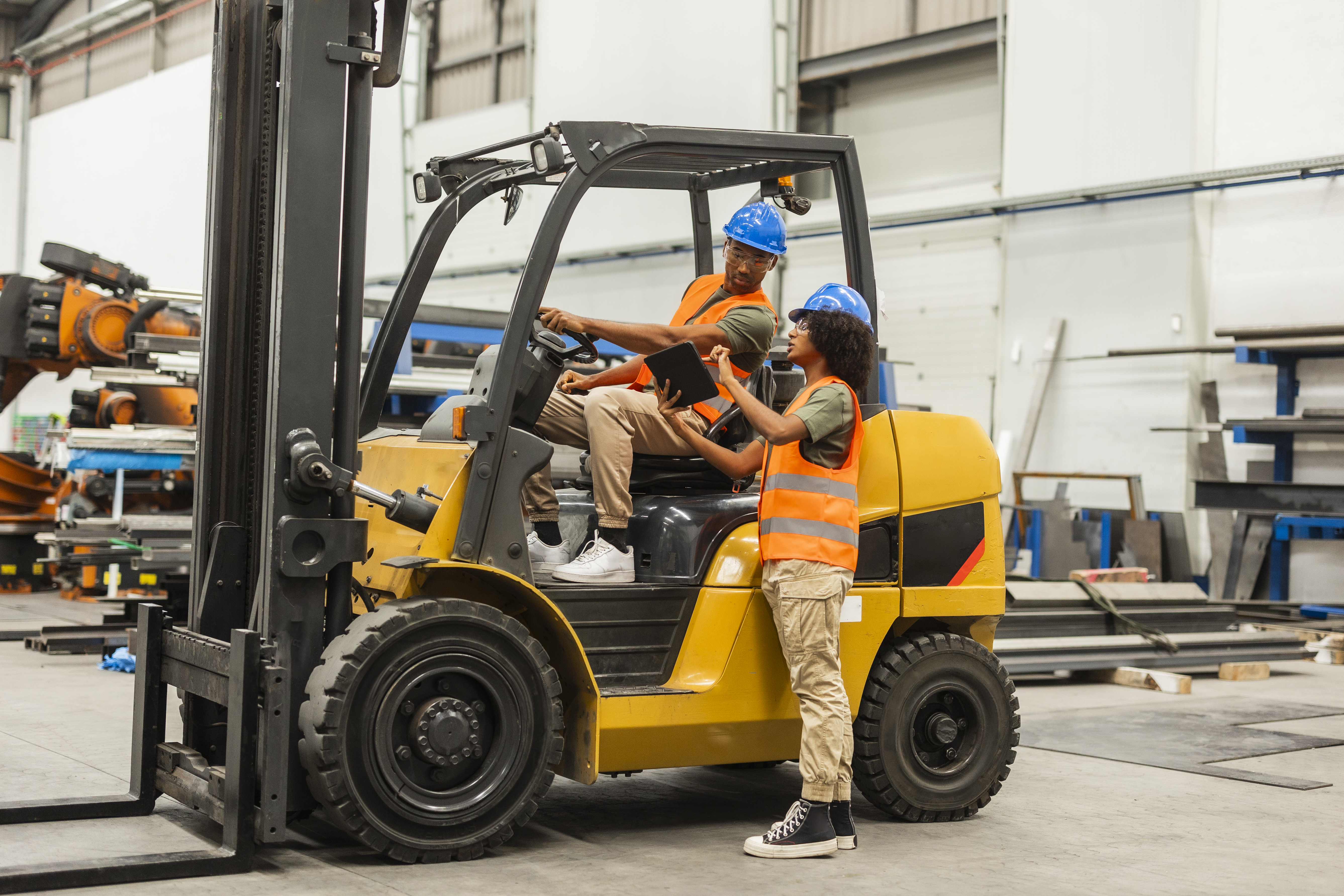An image of two people on a forklift wearing hardhats and orange safety vests.