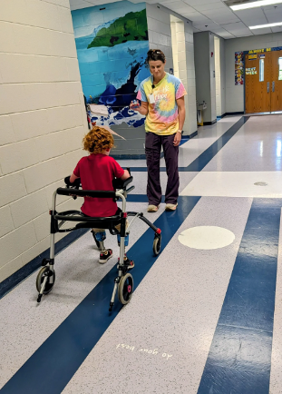 A woman in a tie dye shirt stands in front of a child that has braces on their legs and a walker to help them stand.