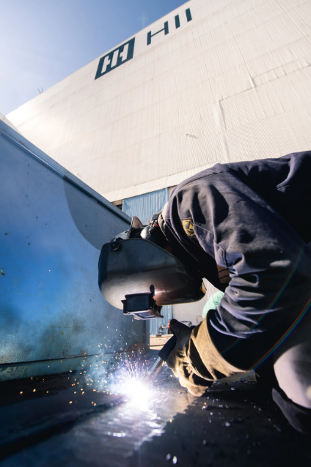 A welder has his protective gear on and a torch in his hands. Sparks are coming out of the torch. The welder is hunched over his project in front of the company sign that reads HII.