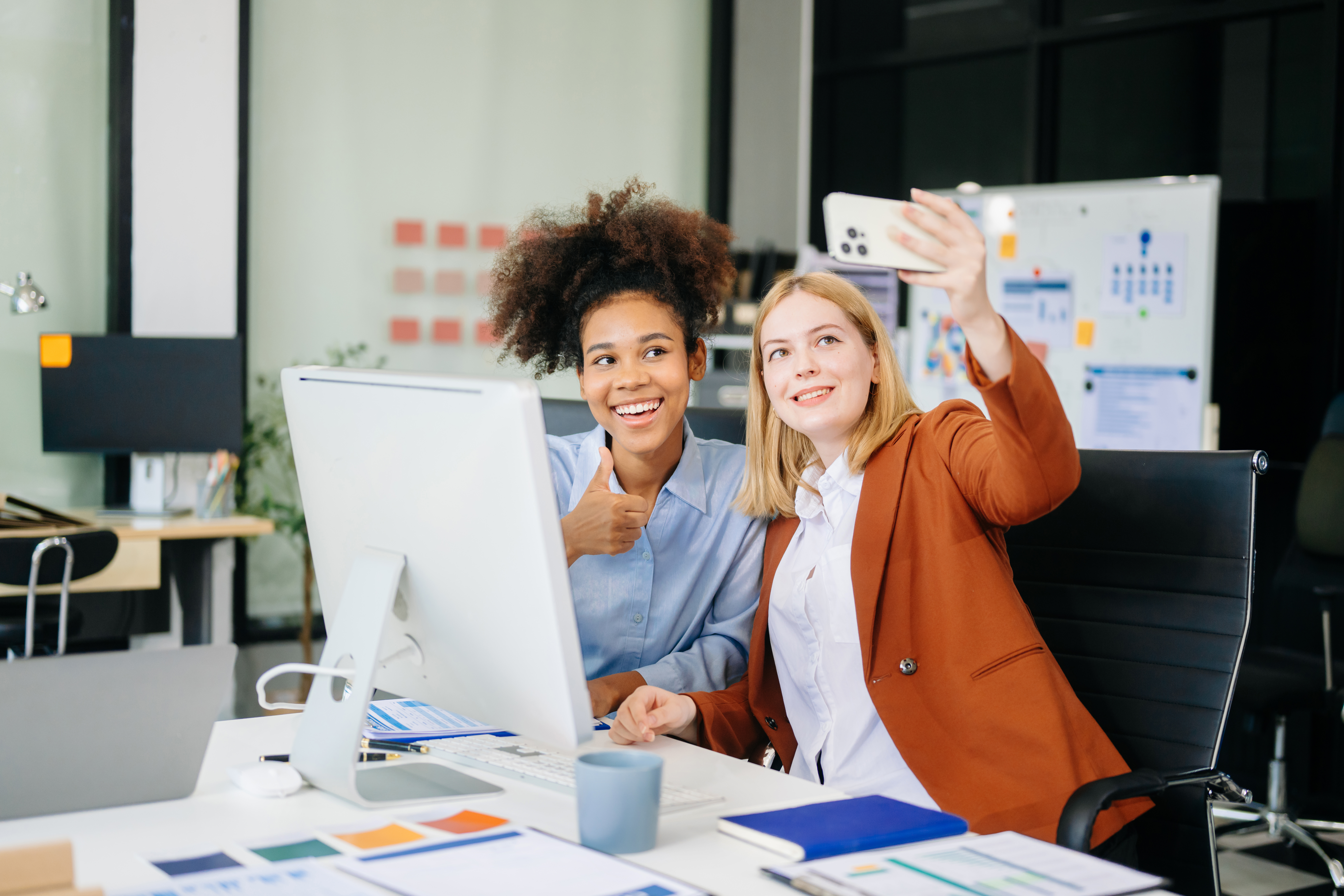 Tracking Social Media Metrics for World Social Media Day. The picture features two women in an office, smiling and holding up a phone.